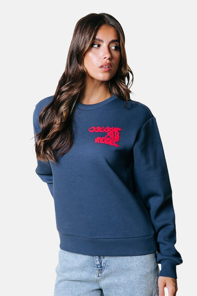 Pull-over bleu de COLOURFUL REBEL avec une impression rouge sur la poitrine, porté avec un pantalon en cuir.