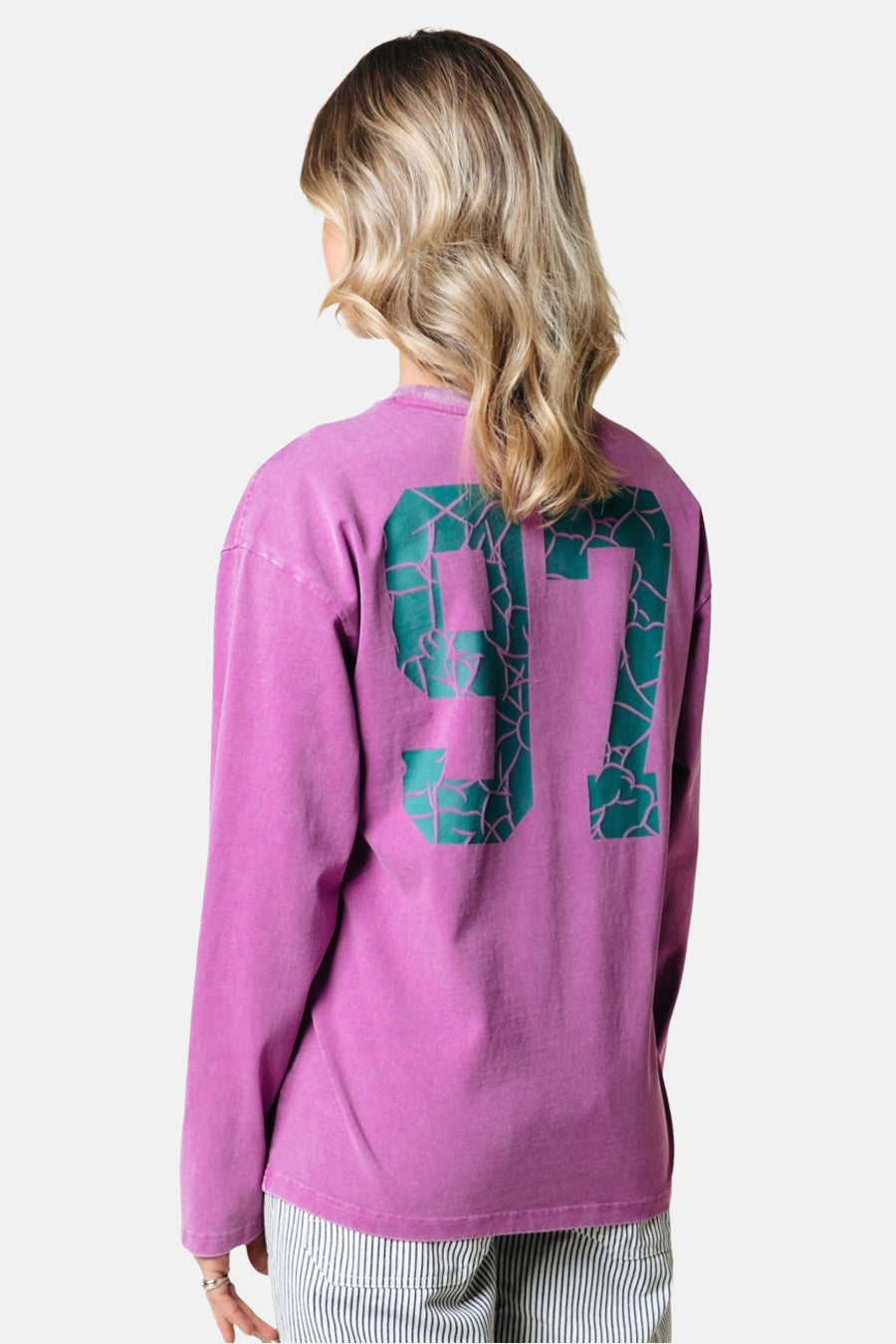 T-shirt rose à manches longues de COLOURFUL REBEL, avec le numéro 97 vert sur le dos, porté avec un pantalon rayé.