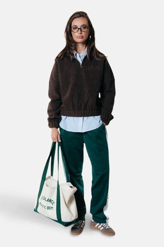 Manteau en laine brune de COLOURFUL REBEL, porté avec un chemisier rayé, des pantalons de jogging verts avec des rayures blanches et une sacoche en toile.