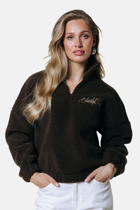 Manteau en laine brune de COLOURFUL REBEL, avec une fermeture éclair courte et un logo brodé, associé à des pantalons blancs et des boucles d'oreilles dorées.