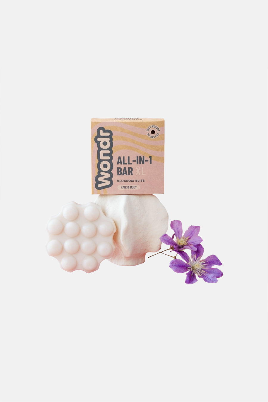 Barre de soin tout-en-un de couleur claire avec relief à picots de WONDR CARE, à côté d'une boîte rose à motifs et de fleurs violettes.