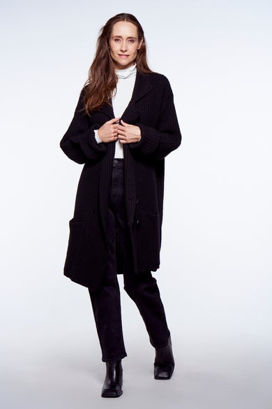 Gilet noir YŪGEN, avec fermeture à boutons et manches longues, porté sur un pull col en V blanc et un pantalon sombre.