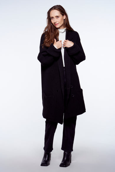 Gilet noir YŪGEN, porté sur un pull col en V blanc et un pantalon noir, avec manches longues et poches à l'avant.