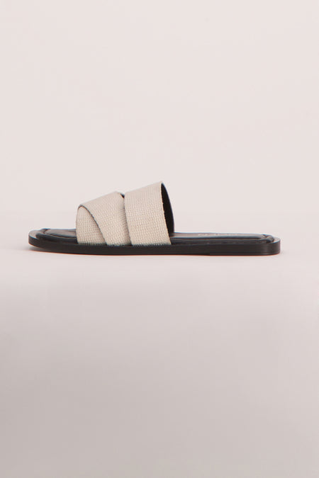 Beige sandalen van YŪGEN, met gekruiste banden over de voet en een platte, zwarte zool.
