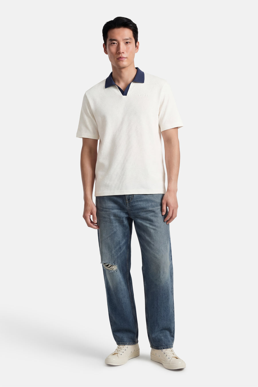 Polo écru à manches courtes de Guess®, avec col bleu, porté avec un jean déchiré et des baskets couleur crème.